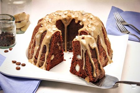 Bolo de café com chocolate. Ele tem um furo no meio, calda de cor bege com raspas de castanha do pará sob prato e com fatia - Metrópoles