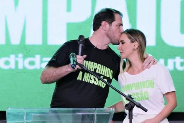 Fotografia colorida de homem com camiseta preto beijando testa de mulher vestida de branço