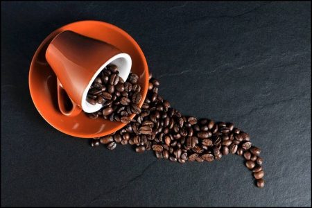 Caneca laranja dentro de tigela da mesma cor com grãos de café saindo sob fundo preto - Metrópoles