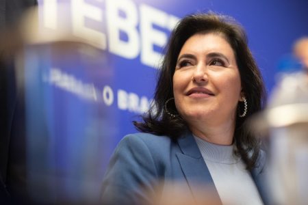 Coletiva de Imprensa da Senadora Simone Tebet, pré-candidata à Presidência da República pelo MDB. Ela tem cabelo escuro e longo, é branca e usa blazer, olhando pro lado em frente a banner azul - Metrópoles