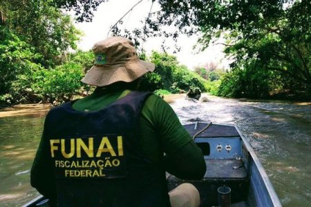 Agente de fiscalização da FUNAI conduz barco em rio no meio de uma mata. Ele usa colete, chapéu e está de costas - Metrópoles