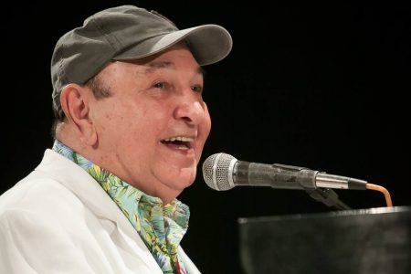 João Donato está de bone preto, com uma camisa colorida e uma jaqueta branca. Ele está cantando e com o microfone à sua frente - Metrópoles