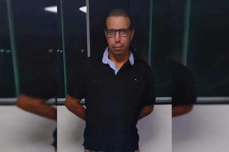 Homem de cabelo curtos e óculos veste camisa polo azul escura e cruza os braços atrás das costas