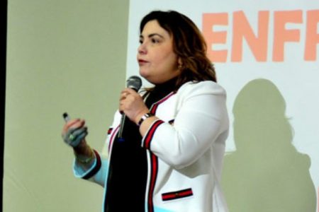 Mulher em pé com microfone nas mãos. Ela usa casaco branco com listra vermelha e camisa preta - Metrópoles