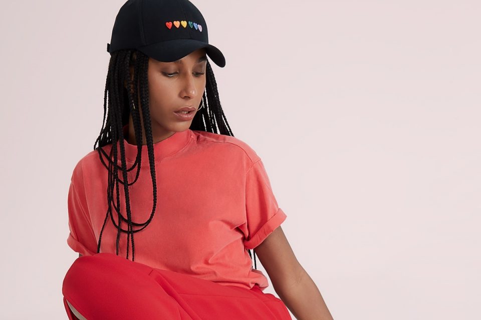 Mulher negra com cabelo trançado posando para foto em um fundo branco. Ela usa uma camiseta vermelha e um boné preto com corações que formam um arco-íris, da bandeira LGBTQIA+. As peças são da Amaro.