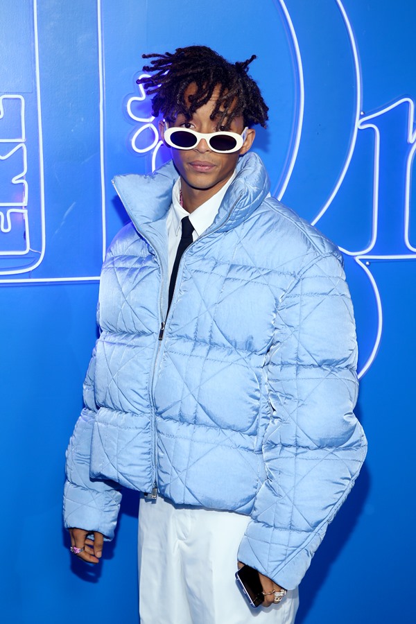Jaden Smith, um jovem rapaz negro com cabelos pretos e de tranças, posando para foto na entrada do desfile da Dior. Ele usa uma camisa de botão branca, gravata preta e um casaco puffer, acolchado, azul, além de óculos escuros com armação branca.