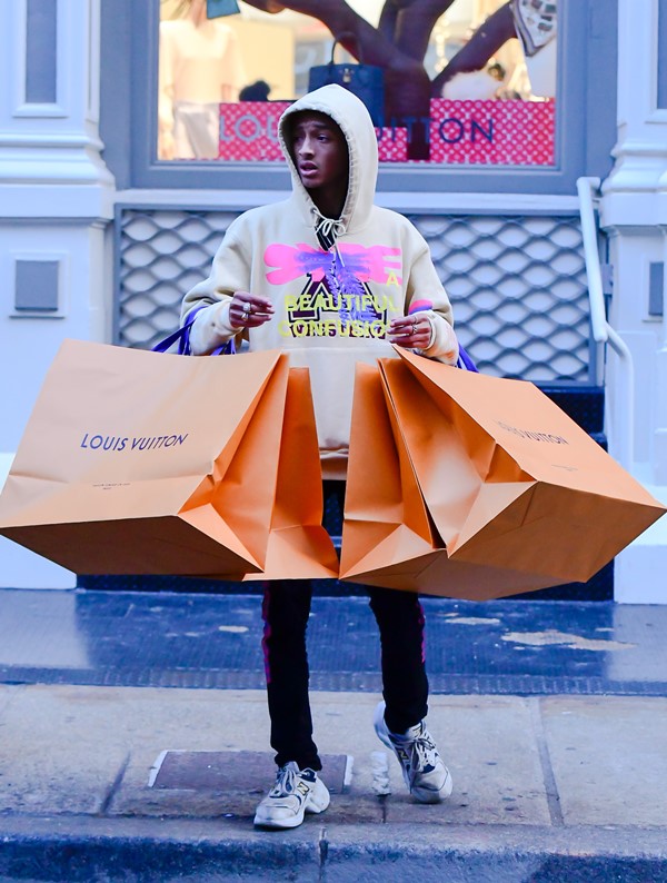 O ator e cantor Jaden Smith, filho de Will Smith, andando pelas ruas de Nova York com sacolas da Louis Vuitton. Ele veste uma calça preta e um moletom bege com estampa rosa e azul na frente.