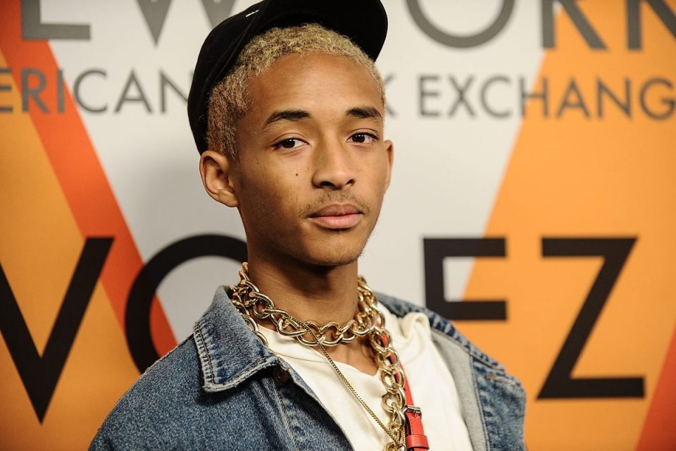 Ator e cantor Jaden Smith, um rapaz negro com cabelo raspado louro, posando para foto em um fundo branco no evento da Louis Vuitton. Ele usa uma camiseta branca, jaqueta jeans e