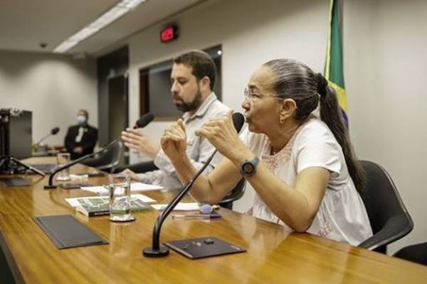 Fotografia colorida de mulher falando em microfone fixado em mesa com homem ao lado