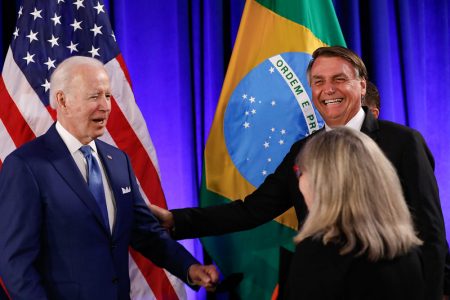 O presidente Bolsonaro cumprimenta o presidente americano Joe Biden na Cúpula das Américas, apertando as mãos enquanto uma mulher loira os olha, de costas. Ao fundo, as bandeiras dos Estados Unidos e Brasil - Metrópoles