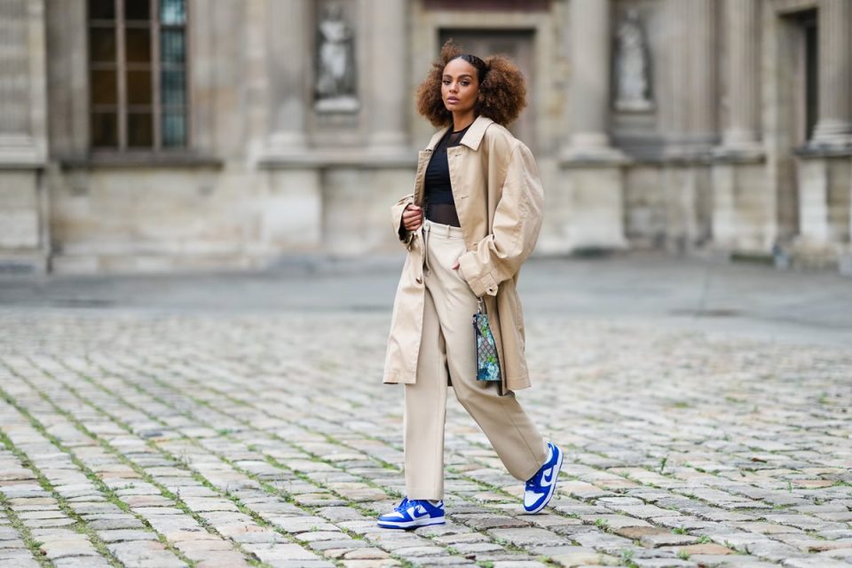Mulher jovem e negra, com cabelo crespo amarrado, andando pelas ruas de Paris durante a semana de moda. Ela usa brincos dourados, camiseta preta transparente de manga comprida, top cropped preto ombro a ombro, casaco bege oversized, calça bege wide leg, bolsa estilo clutch bege com monograma GG marrom e azul com estampa de flores da Gucci, tênis Air Force One de couro brilhante branco e azul royal da Nike.