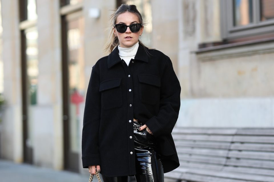 Mulher branca com cabelo liso e longo amarrado em rabo de cavalo. Ela posa para foto nas ruas de Paris. Veste uma blusa branca de gola alta, uma jaqueta preta e uma calça preta de couro. Está de óculos escuros.