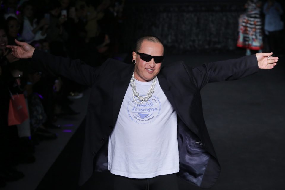 O estilista Walério Araújo, um homem branco de meia idade com o cabelo raspado, na fila final de deu desfile durante a Semana de Moda de São Paulo. Ele usa uma camiseta branca, um blazer preto, colares de pérola e brilhantes e um óculos escuro.