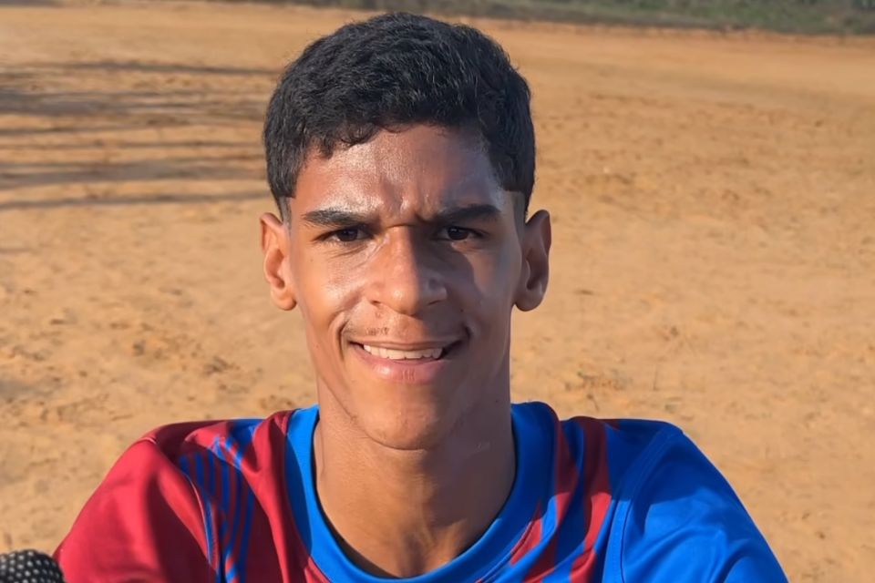 A foto colorida mostra um menino de cabelo castanho vestindo um blusa de time de futebol nas cores vermelho e azul. Ele sorri para a câmera