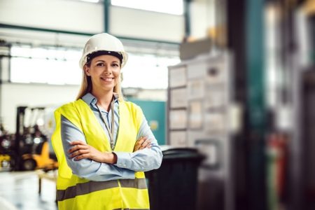 Engenheira vestida com colete amarelo neon e capacete sorri olhando pra câmera em ambiente de trabalho - Metrópoles