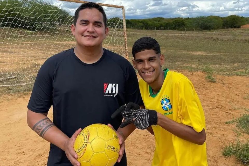 A foto mostra um homem de blusa preta segurando uma bola amarela ao lado de um jovem adulto de blusa amarela de futebol, com luvas de pedreiro, apontando para o homem ao seu lado