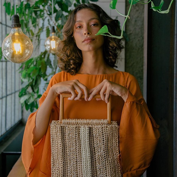 Mulher jovem, morena e com cabelos ondulados longos posando para foto. Ela veste uma blusa de manga longa laranja e segura uma bolsade crochê bege com calça de madeira, no formato retandular.