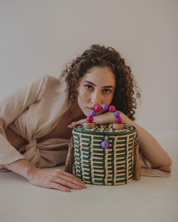 Mulher jovem, branca e com cabelos cacheados longos posando para foto em um fundo branco. Ela veste uma blusa de manga longa bege e segura uma bolsa estilo cesto, de palha, com listras beges e verdes.