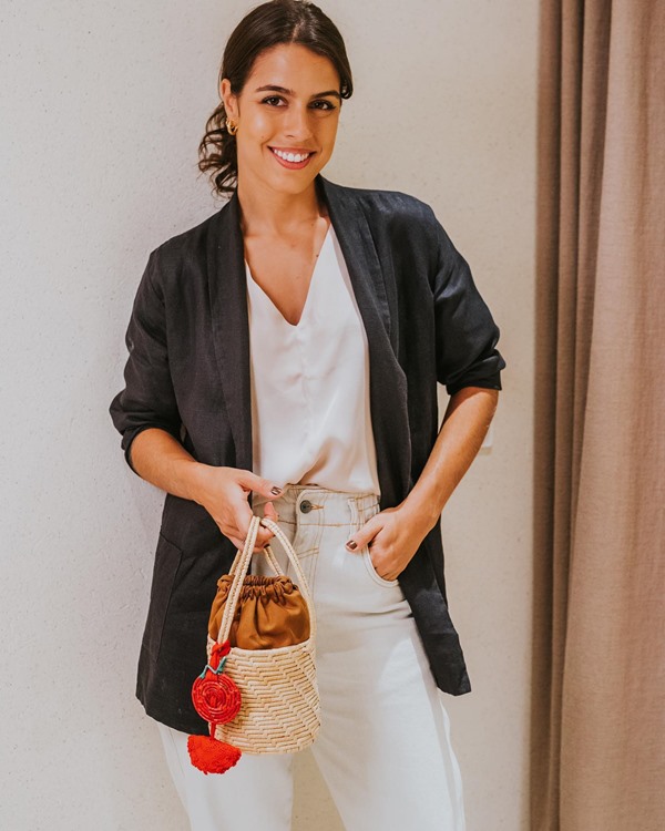 Mulher jovem, morena e com cabelos castanhos amarrados em um rabo posando para foto. Ela veste uma camiseta, blazer preto e uma calça branca. Ela segura uma bolsa de palha com um tecido marrom e um pingente vermelho.