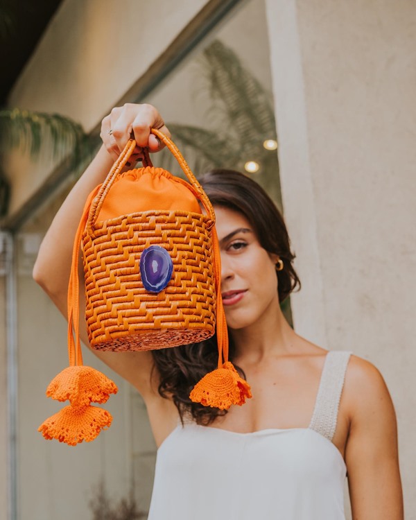 Mulher jovem, morena e com cabelos castanhos ondulados posando para foto. Ela usa um vestido branca de alcinha e segura uma bolsa de palha laranja com um pingente azul.