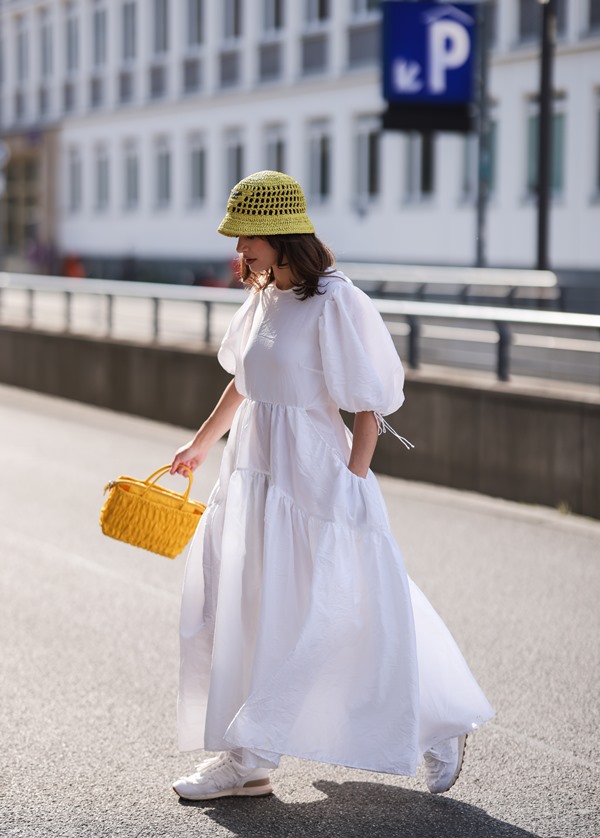 Mulher branca e jovem, com cabelos castanhos lisos e curtos, andando pela rua. Ela usa um vestido branco longo e com mangas bufantes, um chápeu de crochê verde, uma bolsa de palha amarela e tênis esportivos, também brancos.