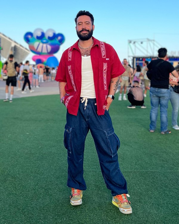 O podcaster e influenciador Samir Duarte posando para foto em um festival de música. Ele é um homem jovem, branco, com barba e cabelos pretos, e veste uma camisa regata branca, camisa social vermelha, calça jeans e tênis cinza com cadarço laranja.