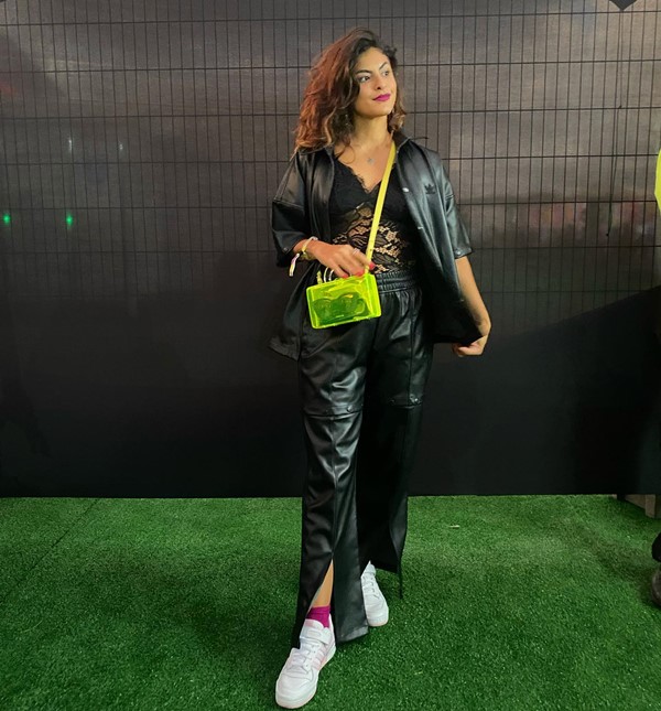 A influenciadora de moda e podcaster Marina Santa Helena posando para foto em um festival de música. Ela é uma mulher jovem e negra, com cabelos ondulados, e veste um maiô de renda, calça e jaqueta de couro, tudo em preto, tênis branco e uma bolsa transpassada verde limão