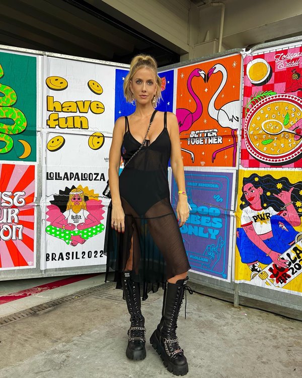 A advogada e apresentadora da CNN, Gabriela Prioli, posando para foto em um mural colorido do festival Lollapalooza. Ela é uma mulher branca e loura e está com o cabelo presto em um rabo de cavalo. Usa um vestido preto transparente e coturnos de couro preto até a altura do joelho.