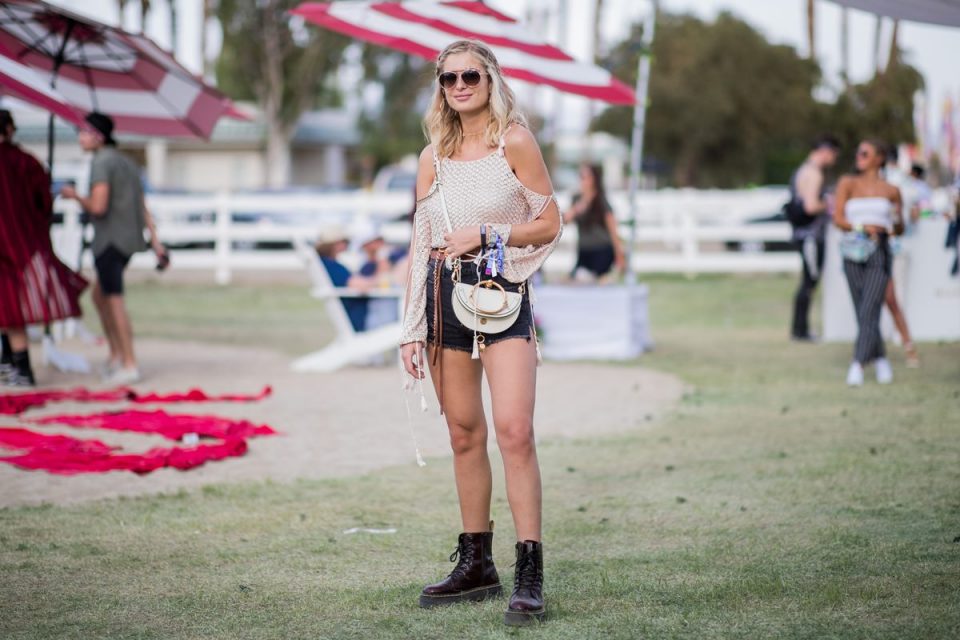 Mulher branca e jovem, com cabelos louros curtos e ondulados, posando para foto no festival de música Coachella, nos Estados Unidos. Ela está de óculos escuros e usa uma blusa bege com recorte ombro a ombro, um shorts jeans preto, um coturno de couro preto e uma bolsa transpassada branca da marca Chloé.