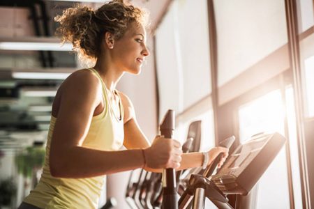 Mulher parda se exercitando em máquina de musculação na academia. Ela aparece de lado, com roupas de malhação, e sorri - Metrópoles