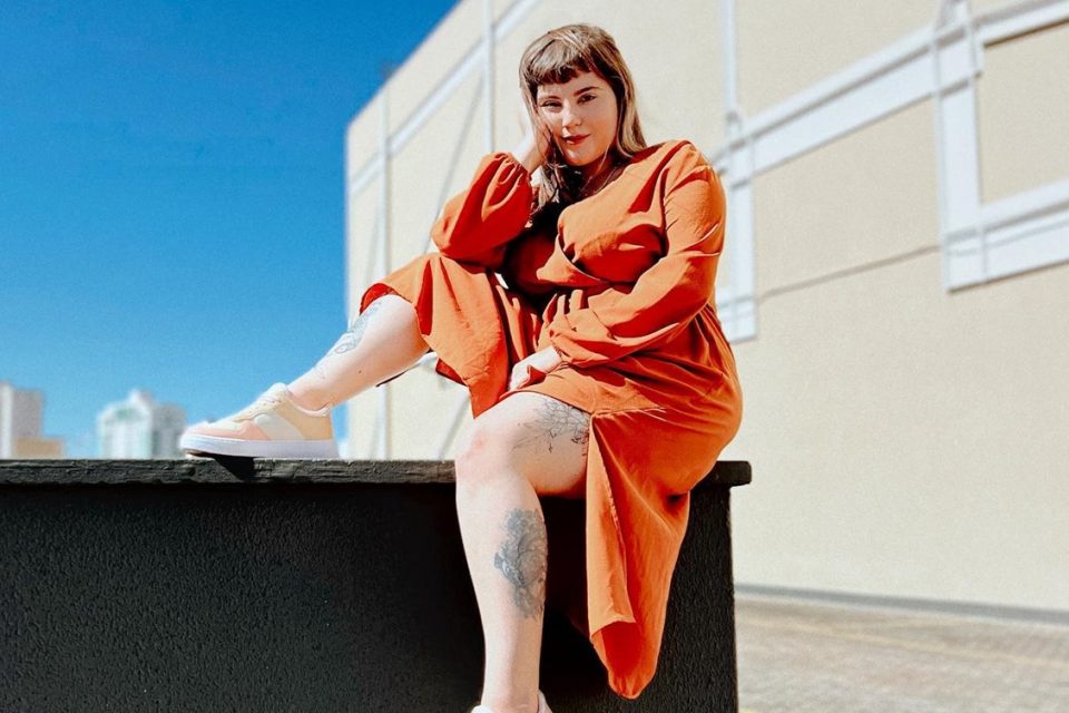 A influenciadora digital brasiliense Nanna Fernandes, uma mulher branca, jovem, com cabelos lisos e com tatuagens pelo corpo. Ela está posando para foto em um muro e usa um vestido laranja e um tênis branco.