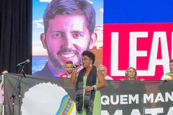 Fotografia colorida de mulher discursando com foto de homem branco ao fundo