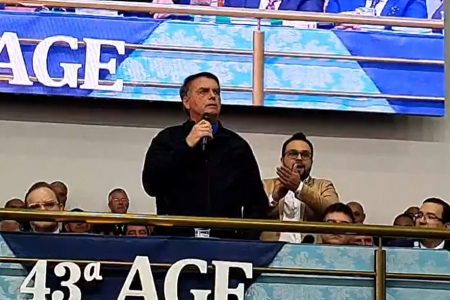 Em Juiz de Fora (MG), o presidente Bolsonaro fala sobre facada que levou na cidade em 2018. Ele discursa em mezanino cercado de outras pessoas com microfone na mão e jaqueta preta - Metrópoles