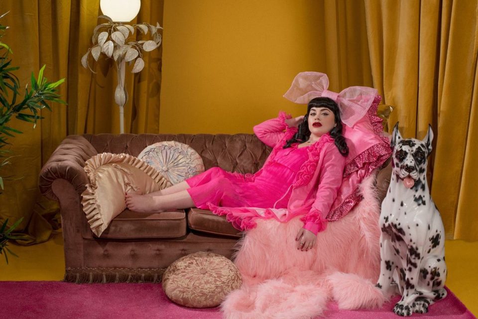 Mulher com deficiência em editorial de moda: ela usa look rosa e está deitada em um sofá, com cachorro ao lado