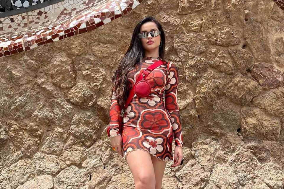 Em frente a um muro de pedras. Juliette usa vestido transparente estampado, feito de tule, com flores em vermelho e laranja