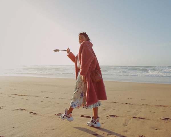 A atriz Bruna Marquezine posando para foto na praia. Ela é uma mulher jovem, branca e de cabelos curtos e lisos, e usa uma saia, um casaco de pelo rosa e um tênis branco e preto