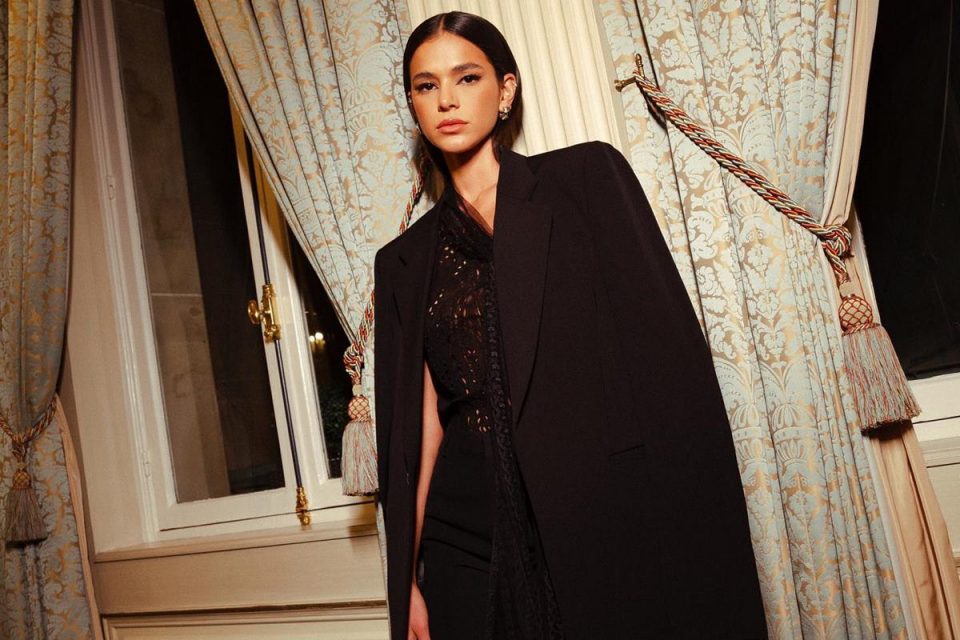 A atriz Bruna Marquezine posando para foto na janela de um hotel em Paris. Ela usa um camisa de botão com renda, um blazer e uma calça. Todas as peças são pretas.
