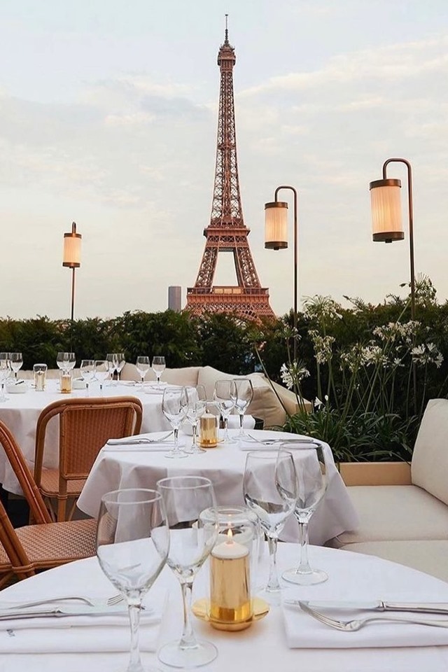 Foto colorida. Ao fundo, aparece a Torre Eiffel. Mesas e cadeiras