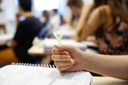 Pessoa escrevendo em caderno em primeiro plano. Em segundo plano, outras pessoas estudam em uma sala de aula concurso - Metrópoles