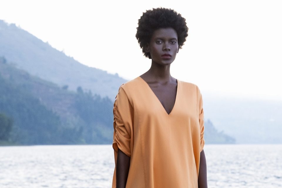 Mulher negra e jovem, de cabelo curto e cacheado, posando para foto em uma praia, em cima de uma jangada. Ela usa um vestido longo marrom