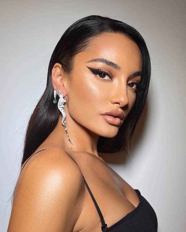 A estilista Amina Muaddi, uma mulher jovem de pele morena e cabelo liso longo, posando para foto em um fundo branco com delineado gatinho preto