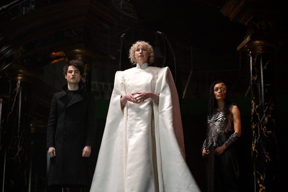 Série Sandman. Na imagem, mulher ao centro usa roupa branco com ombros marcados e caimento lateral, ao lado de homem com roupa preta e mulher com look prateado