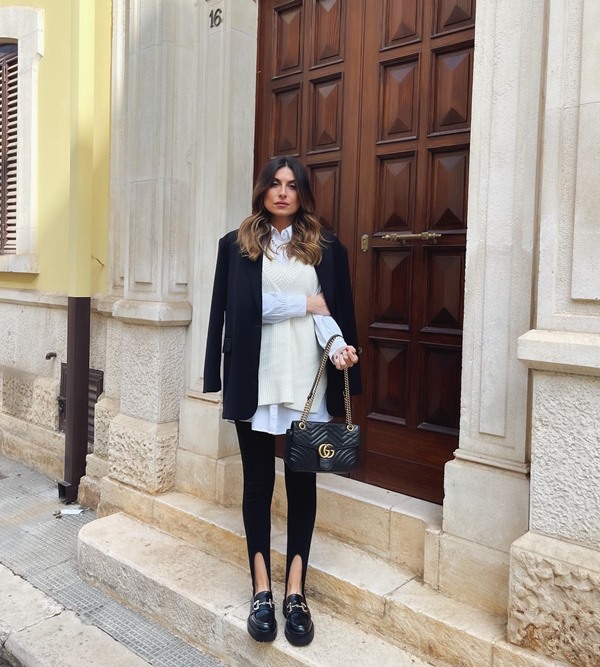 Mulher branca e jovem, de cabelo liso médio, posando para foto nas ruas de Paris, em frente a uma porta de madeira antiga. Ela usa uma camiseta branca, colete bege, blazer preto, legging preta, sapato mocassim e bolsa de couro, também preta, da marca Gucci