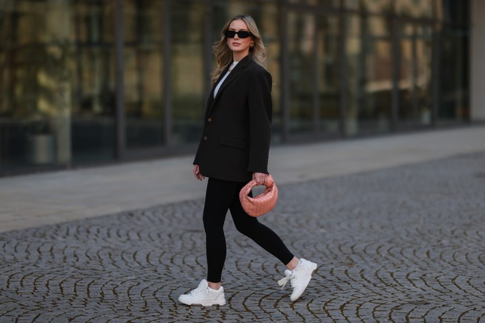 Mulher branca e jovem, de cabelos loiros longos e ondulados, posando para foto na rua. Ela usa uma blusa branca, blazer preto, calça legging preta, tênis esportivo branco e uma bolsa de mão de couro rosa, da marca de luxo Bottega Veneta