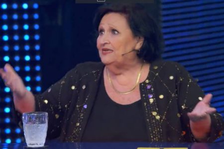 Foto colorida de Déa Lúcia usando roupa preta brilhante e microfone no Domingão - Metrópoles