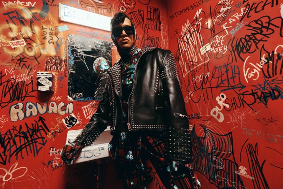 Fotos de campanha da coleção da Tommy Hilfiger com o estilista Richard Quinn. Um homem jovem e de pele branca está em um banheiro vermelho e usa uma roupa xadrez preta e vermelha e uma jaqueta de couro.