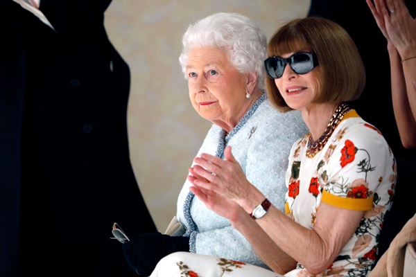 A Rainha Elizabeth II sentada ao lado de Anna Wintour, jornalista de moda da revista Vogue, durante a Semana de Moda de Londres em 2018. A monarca usa um casaco azul claro de tweed e a jornalista está com um vestido branco com flores vermelhas e amarelas e um óculos escuro.