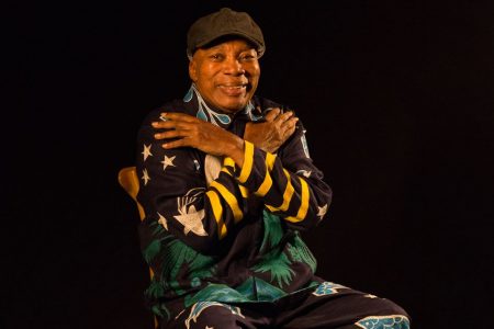 Foto colorida de Milton Nascimento. Ele é um homem negro, está sentado, usa boina preta e uma roupa preta com detalhes coloridos. O fundo é preto - Metrópoles