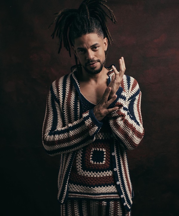 O ator Paulo Lessa em ensaio de fotos com um fundo escuro. Ele é um homem jovem, negro e com dreads, e veste um conjunto de camisa, regata e calça da marca baiana Ateliê Mão de Mãe. As peças são feitas de crochê nas cores branca, azul e vermelho