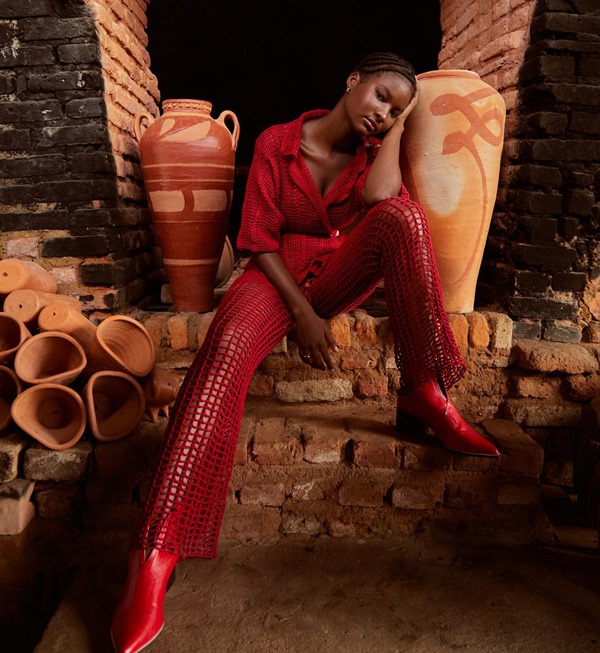 Modelo jovem e negra posando para foto em meio a vasos de barros em estúdio de artesanato na Bahia. Ela usa uma camisa e uma calça, ambas as peças confeccionadas em crochê vermelho. A foto é de campanha da marca Ateliê Mão de Mãe.